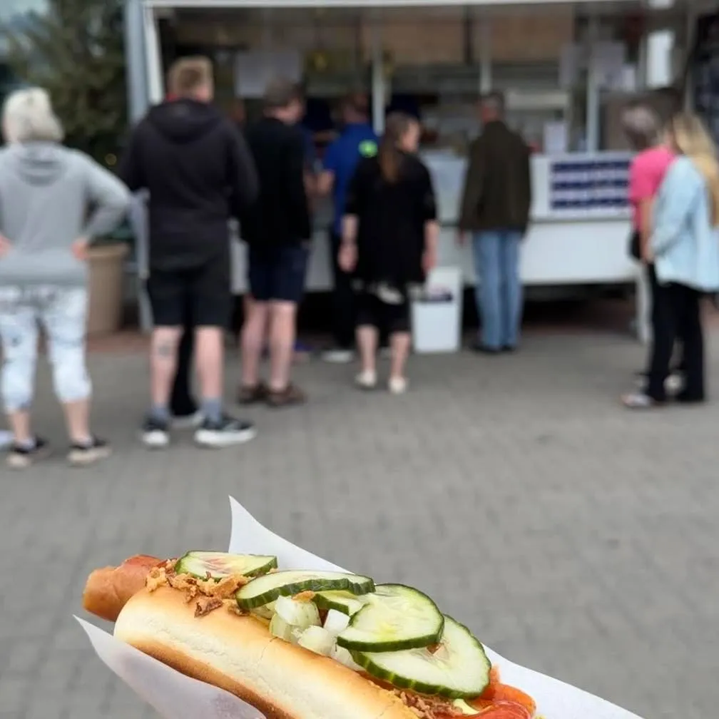 Gæster i kø ved pølsevognen med en nylavet hotdog i forgrunden